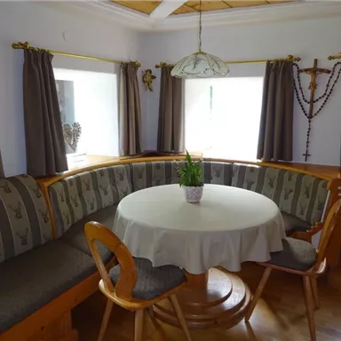 A cozy dining area with a round table and a bench. The windows are decorated with curtains, and a plant decoration is in the middle of the table.