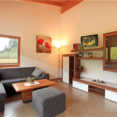 A modern living room with a gray couch, a wooden coffee table, and a matching stool. There are windows and some decorative elements on the wall.
