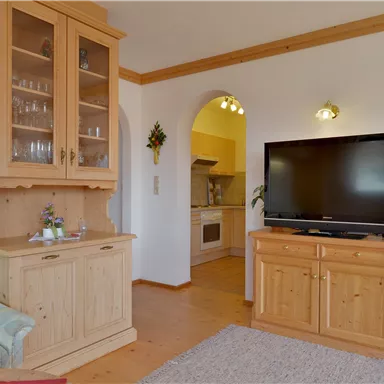 A cozy living room with light wood and a television. In the background, the open kitchen is visible.