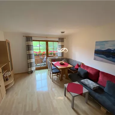 Ein gemütliches Wohnzimmer mit einer Couch, einem Tisch und einem Fernseher. Große Fenster mit Blick auf einen Balkon und blühende Pflanzen.