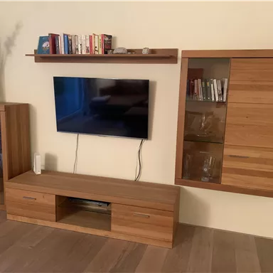A modern living space with a television on the wall and a wooden shelf underneath. Next to the television is an elegant cabinet with glass doors.