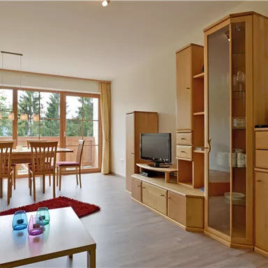 A bright living room with wooden furniture and large windows. In the middle stands a table with colorful glasses and a red carpet.