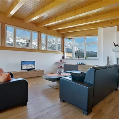 A modern living room with two black sofas and a wooden ceiling. Large windows offer a view of mountainous landscapes.