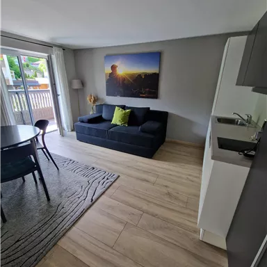 Ein modernes Wohnzimmer mit einem dunkelblauen Sofa und einem hellen Tisch. An der Wand hängt ein Bild mit einer Landschaft und einem Sonnenuntergang.