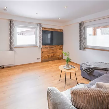 A bright living room with a cozy sofa and a small table. The walls are painted in neutral colors, and large windows let in plenty of light.