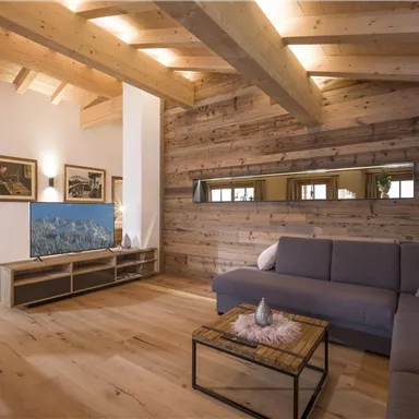 Ein modernes Wohnzimmer mit Holzdecke und -boden. Eine bequeme Couch steht vor einem TV-Möbel, das einen Fernseher zeigt.