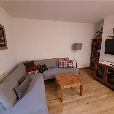 A cozy living room with a gray sofa, a wooden table, and a television on the wall. The decor is modern and warm, with wood and textile decorations.