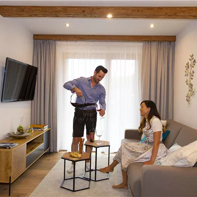 Ein gemütlicher Wohnraum mit einem Mann, der Wein einschenkt, und einer Frau, die entspannt auf der Couch sitzt. Der Raum ist modern und hell mit Holzdetails.