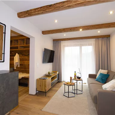 Ein gemütliches Wohnzimmer mit Holzdeckenbalken und moderner Einrichtung. Ein Sofa, ein Couchtisch und große Fenster sorgen für eine helle, einladende Atmosphäre.