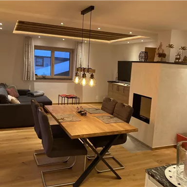 A modern living room with a dining area and a cozy atmosphere. Large windows provide plenty of light and a view outside.