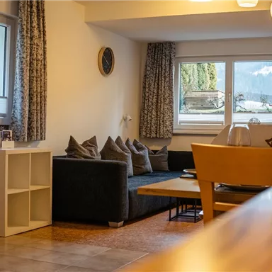 A cozy living room with a gray sofa and a table.  
Large windows offer a view of the nature outside.