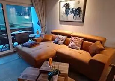 A cozy living room with a brown sofa and a wooden table. Large windows offer a view of the surrounding landscape.