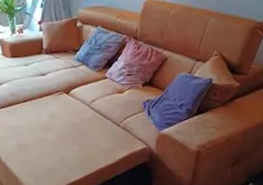 A cozy, orange sofa with colorful cushions is in a bright room. In the background, windows and a table are visible.
