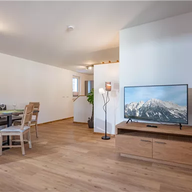 Ein modernes Wohnzimmer mit Holzfußboden und gemütlicher Einrichtung. Ein Fernseher steht auf einem Holzschrank, während ein Esstisch mit Stühlen im Hintergrund zu sehen ist.