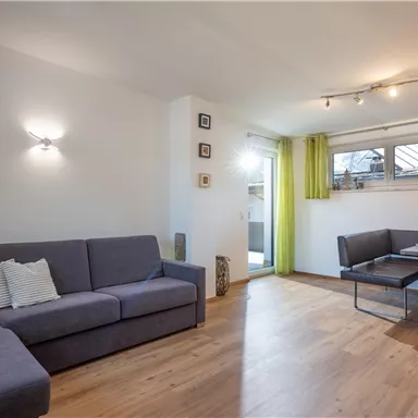 Ein modernes Wohnzimmer mit einer grauen Couch und einem Tisch für vier Personen. Große Fenster mit grünen Vorhängen lassen viel Licht herein.