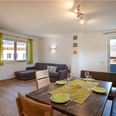 Ein gemütliches Wohnzimmer mit einer Sitzecke und einem Esstisch. Die Wände sind hell, und grüne Vorhänge sorgen für eine freundliche Atmosphäre.