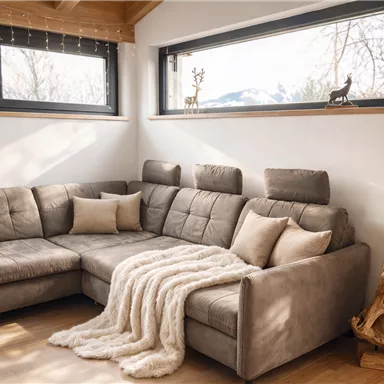 Ein modernes Wohnzimmer mit einem beigen Sofa und einer gemütlichen Decke. Große Fenster bieten einen Blick auf die Natur draußen.