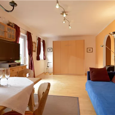 Ein gemütlicher Wohnraum mit blauen Sofa und Esstisch. Große Fenster lassen viel Licht herein und verleihen dem Raum eine einladende Atmosphäre.