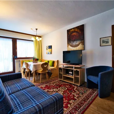 Ein gemütliches Wohnzimmer mit einer blauen Couch, einem Tisch und Stühlen. Der Raum hat große Fenster mit leichtem Tageslicht und eine moderne Fernsehecke.