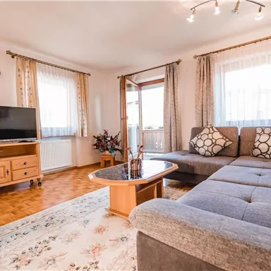 A bright living room with a gray sofa and a coffee table. On the other side, there is a television on a wooden cabinet, and large windows provide plenty of daylight.
