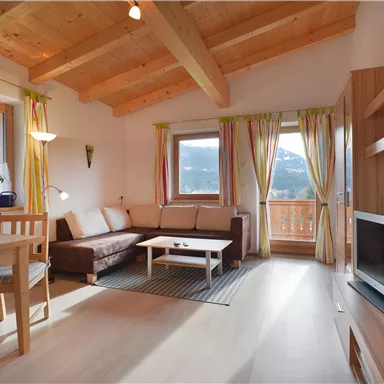 A cozy living room with a wooden beam ceiling and large windows. The room is bright and cheerfully furnished, with a sofa, table, and plants.