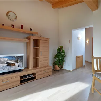 A bright, modern living room with a television, a wooden shelf wall, and a table with chairs. A green plant accent brings life to the room.