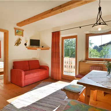 A cozy living room with a red sofa and dining table. Large windows offer a view of nature and let in plenty of light.