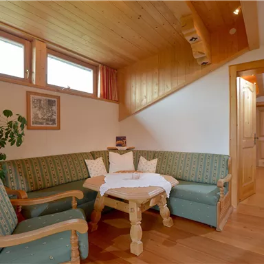 Ein gemütliches Wohnzimmer mit einer grünen Sitzecke und Holzelementen. Im Hintergrund ist ein geschlossener Raum mit einem Bett sichtbar.