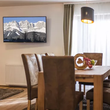 An inviting dining area with a wooden table and cozy chairs. A television hangs on the wall featuring a picture of snow-capped mountains.