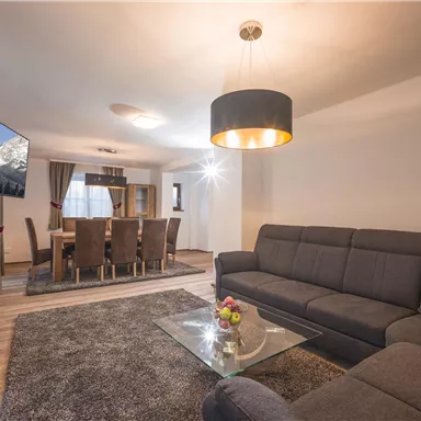 A modern living room with a gray sofa and a glass table. In the background, there is a dining table with chairs and a television.