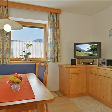 Ein gemütlicher Wohnraum mit einem Holztisch und bunten Vorhängen. Im Hintergrund steht ein Fernseher und eine Fensterbank mit Pflanzen.