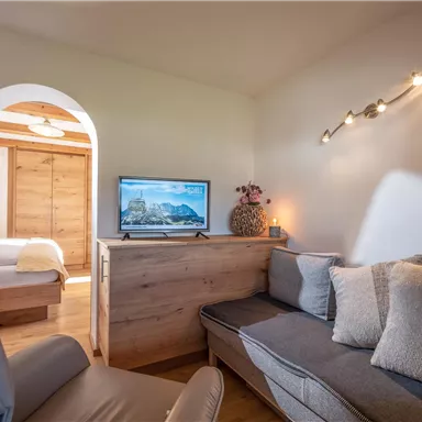 Ein gemütlicher Raum mit einem grauen Sofa und einem Fernseher. Der Raum ist hell und modern eingerichtet und hat einen Zugang zu einem weiteren Bereich.