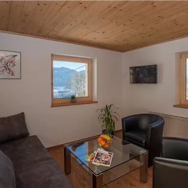Ein gemütliches Wohnzimmer mit einer grauen Couch, zwei schwarzen Stühlen und einem Glastisch. Große Fenster bieten einen Blick auf die Berge und viel Tageslicht.