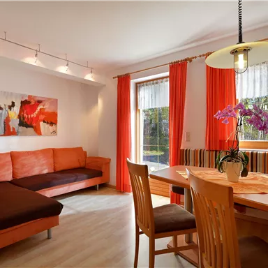 A modern living room with an orange couch and a dining area. Large windows let in plenty of daylight, and a few plants beautify the space.