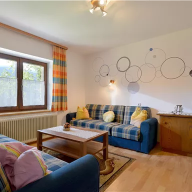 A cozy living room with blue sofas and colorful cushions. Large windows let in plenty of light, and a wooden cabinet complements the decor.