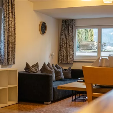Ein gemütliches Wohnzimmer mit einem dunkelgrauen Sofa und großen Fenstern. Der Raum ist hell und modern eingerichtet.