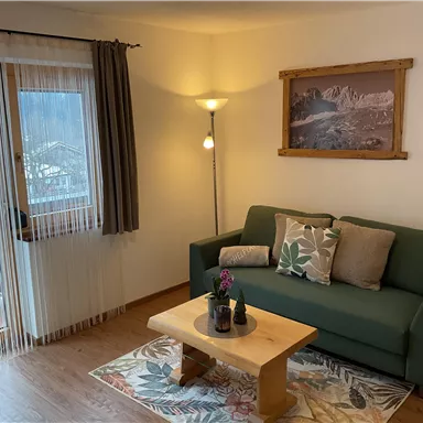 An inviting living room with a green sofa and a wooden table. Bright light shines through the windows, creating a cozy atmosphere.