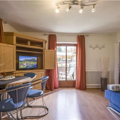 A bright living room with a blue couch and a dining table. Large windows offer a view of an outdoor area.