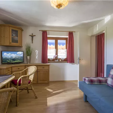 A cozy living room with a blue couch and a wooden table. The windows are decorated with red curtains and offer a view of the landscape.