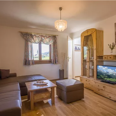A cozy living room with a large sofa and wooden shelves. A TV displays a landscape, while the light gently shines from a chandelier.