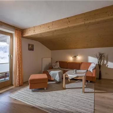 Ein gemütliches Wohnzimmer mit einem orangefarbenen Sofa und einem kleinen Tisch. Große Fenster bieten einen Blick auf die Berge und das Tageslicht kommt hinein.