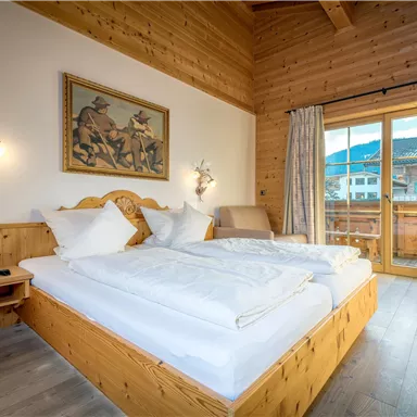 A cozy bedroom with wooden walls and large windows. The room features a double bed and a balcony with a view of the mountains.