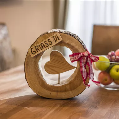 A wooden decoration with the inscription "GRIASS DI" and a heart in the middle stands on a table. In the background, a bowl with various fruits can be seen.