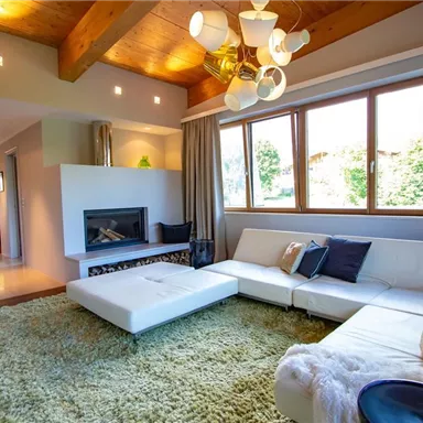 Ein modernes Wohnzimmer mit einem weißen Sofa und einem gemütlichen Teppich. Große Fenster lassen viel Licht hereinfällen und bieten Ausblick auf die Natur.
