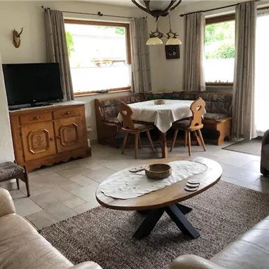 A cozy living room with a large table and wooden furniture. The windows let in a lot of daylight and the room feels inviting.