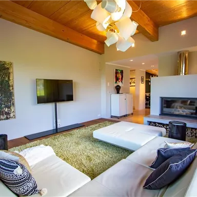 Ein modernes Wohnzimmer mit einem weißen Sofa, einem Fernseher und einem Kamin. Der Raum hat große Fenster und einen warmen Holzdecken.