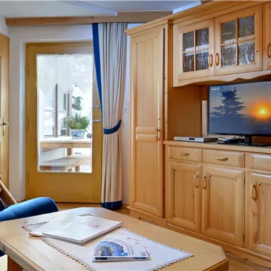 A cozy living space with a blue armchair, a wooden furniture wall, and a television. A beautiful view of the outdoors is visible through the door.
