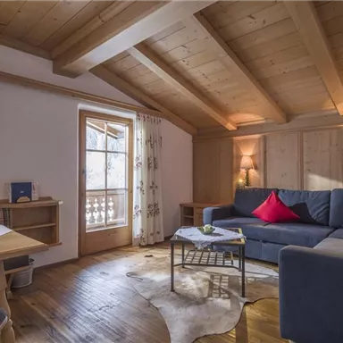 Ein gemütliches Wohnzimmer mit Holzdecke und großen Fenstern. Es gibt ein Sofa mit roten Kissen und einen Esstisch.