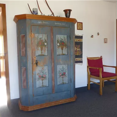 Ein traditioneller Schrank mit bunten Blumenmotiven steht in einem hellen Eingangsbereich. Neben dem Schrank befindet sich ein roter Stuhl aus Holz.