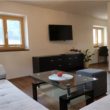 A modern living room with a white sofa, a glass table, and a TV on the wall. Large windows let in a lot of natural light.